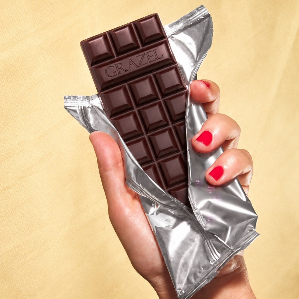 A hand with red nail polish holds a dark chocolate bar partially unwrapped from shiny silver foil against a warm beige background. The chocolate bar is embossed with the brand name GRAZEL.