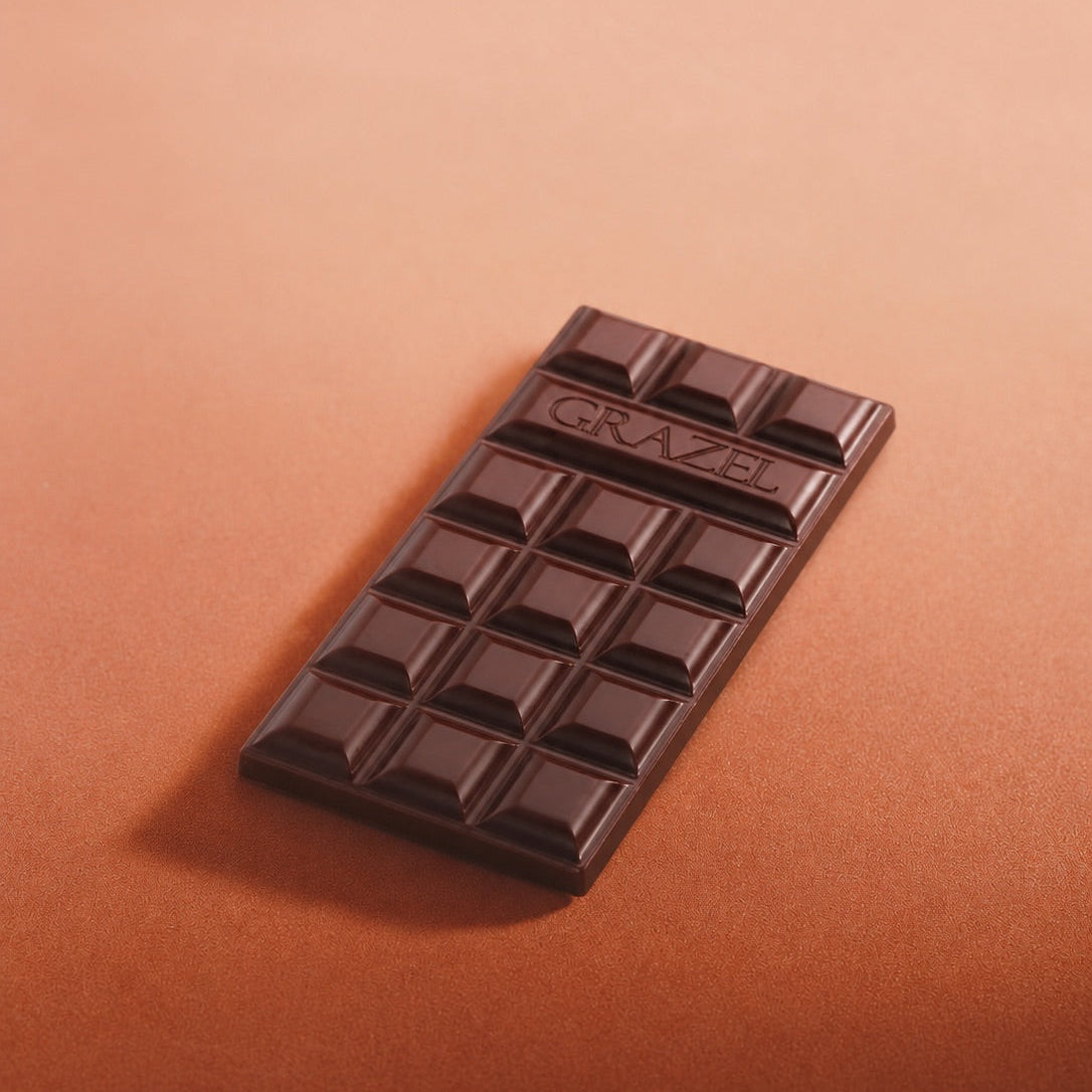A dark chocolate bar with the brand name GRAZEL embossed on the top, resting diagonally on a warm, solid brown background.